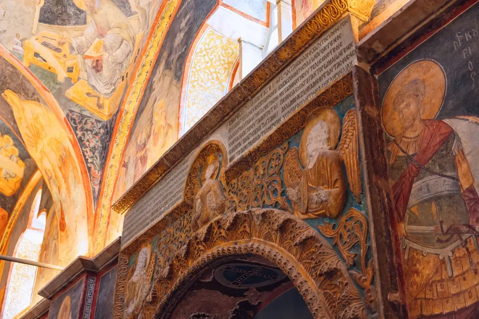 Carved stone arch with busts of angels and a long Greek inscription above it, next to a fresco of a saint in armour